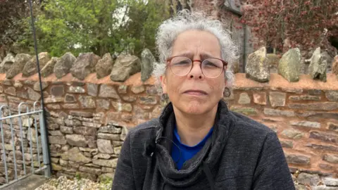BBC Vik Fielder outside. She is wearing glasses, has curly grey hair and is in a dark grey jacket over a blue T-shirt. She is in front of a stone wall. 
