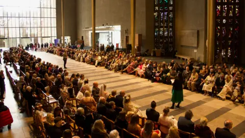 The central area of Coventry Cathedral with people sat in long rows facing each other and two presenters stood in the middle