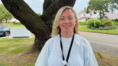 A woman standing in front of a tree, wearing a white T-shirt and a black lanyard, smiling.