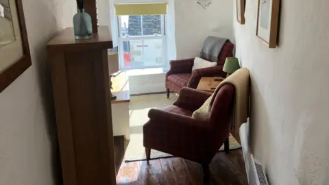 Tiny living room with two chairs, narrow shelving and a window through which the house opposite is visible.