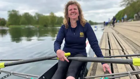 EMMA BAUGH/BBC Kate Lindgren wearing a blue fleece sitting in a rowing boat holding her oars. Her left hand is leaning on wooden decking, and in the left of the photo and in the background is the river and some trees on the bank.