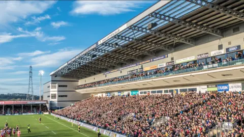 A packed rugby stadium with thousands of spectators seated in a large modern stand on a sunny day. Players gather on the pitch in the foreground, while advertising boards line the stand and a tall electricity pylon stands in the background against a blue sky.