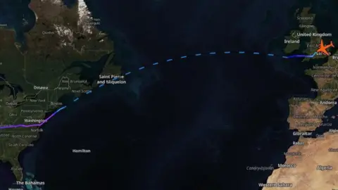ADS-B Exchange Satellite map showing the United States, Atlantic Ocean and Europe. The sea is dark blue and the land green and brown in colour. There is a purple and blue line indicating a flightpath from the US to the UK.