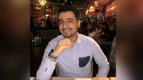 Family photo Rustam, a young man sitting in a restaurant wearing a smart blue shirt and smiling at the camera.