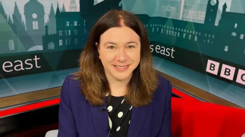 Nic Rigby/BBC A woman sits in a BBC TV studio, smiling for the camera. She has shoulder-length hair and is wearing a black and white spotted dress and a blue jacket.