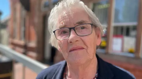 An 86-year-old woman with grey hair wears dark glasses and a dark top is looking straight at the camera.