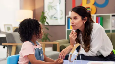 Getty Images The picture shows a female speech therapist with long brown hair pointing her index finger to her lips to demonstrate mouth movements.while sat a low table opposite a young girl. 