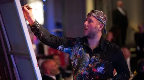 Trivandi A man with a backwards cap painting, his shirt is covered in paint and he is looking at a canvas holding a paintbrush in his right hand. 