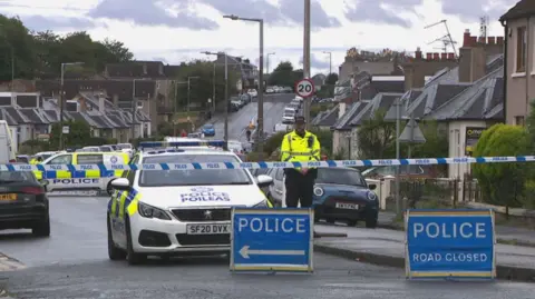 A residential street in Edinburgh taped off with police tape. There are blue signs that read police road closed and several police cars and vans behind the tape while one officer stands guard.
