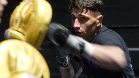 Williams is sparring in the ring, hitting yellow focus pads worn by a trainer