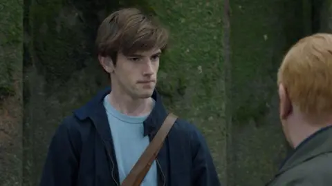 Jude stands in front of a mossy wall, facing another character whose face we can't see. It is Detective Inspector Max Arnold, played by Adrian Scarborough, who has ginger hair and wears a green coat. Jude wears a blue t-shirt and navy collared jacket, with a brown satchel strap across his body. He had brown hair swept over to one side. 