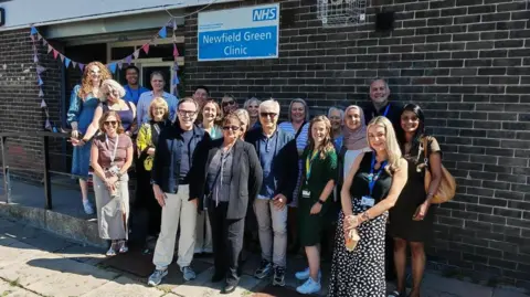 A photo of members of staff gathered outside the Newfield Green Clinic, which has a colourful banner draped around its entrance.