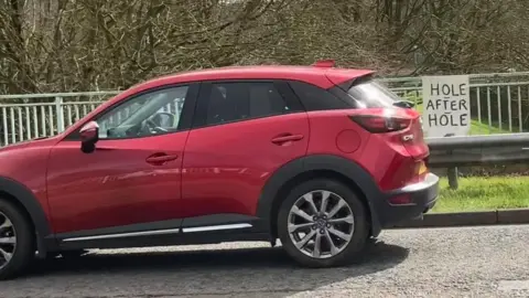 Martin Heath/BBC A red car passes a "hole after hole" sign on railings on a roundabout