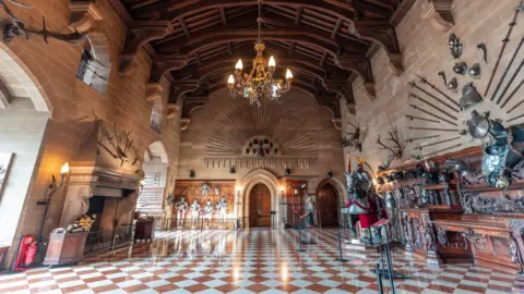 Warwick Castle A grand hall of a castle, with collections of armour at eye-level and a very high carved wooden ceiling. Sandstone coats the wall and chequered white and brown tiles on the floors.