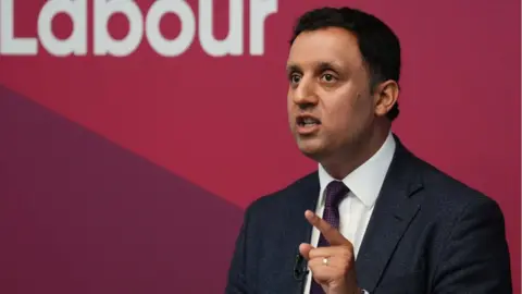 PA Media Anas Sarwar, a man with dark hair, speaks while standing in front of a red Labour sign. He is wearing a dark suit, white shirt and purple tie, and pointing with his left hand.