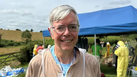 Brian has short grey hair and is smiling at the camera as he wears glasses and a beige coloured beekeeping suit.