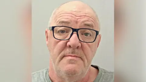 Police custody image of David Berry with receding grey hair and grey beard wearing black glasses and grey t-shirt. 