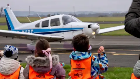 Viv Porteous Children in high-vis vests watching a light aeroplane take off