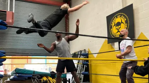 The character Demetrius flies in the air after jumping from the ringside, ready to tackle the character Lysander. This takes place inside a wrestling ring.