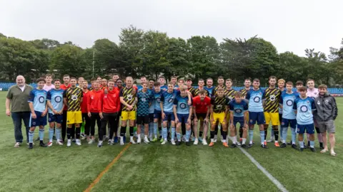 Hannah McHugh/FC Isle of Man Dozens of males in different coloured kits stand on a green pitch with their arms around each other.
