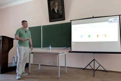 Sergiy Sylantyev Sergiy Sylantyev standing in a lecture hall looking at a screen, he is wearing beige trousers and a green t-shirt and is holding a controller.