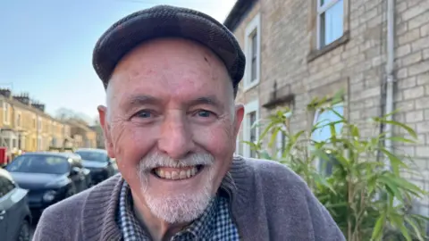 An image of Bob Asworth, who is standing on the pavement outside his house. There is a bush to his right and cars parked to his left. He is smiling. He is an older man with a white beard and is wearing a flat cap. He is also wearing a checked shirt with a collar and a grey cardigan.
