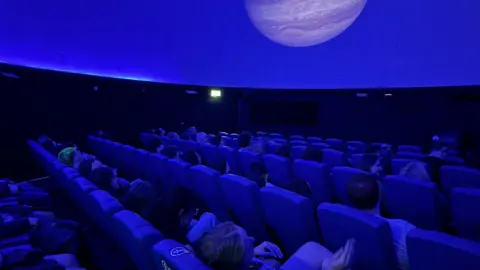 The audience sit in rows of seats in the planetarium's main dome room. The room is lit blue.