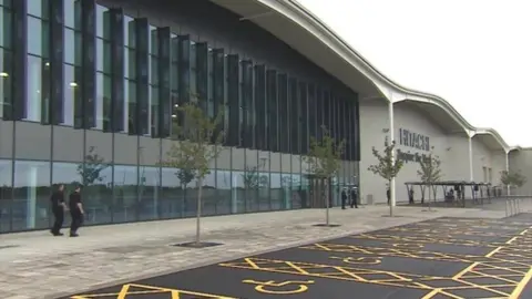 The Hitachi factory. Two men walk pass the glass fronted building. The Hitachi logo can be seen high up on a concrete wall to the right. Young trees are planted by the pavement and next to disabled parking bays.