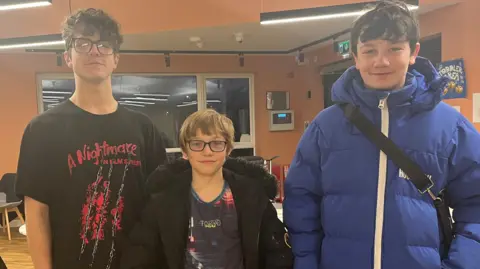 Thomas has brown curly hair and glasses. He is wearing a black shirt with red writing on it advertising the film 'A Nightmare on Elm Street', Vinny is in the middle and wears a black puffer jacket and glasses and Joe is to the right wearing a blue puffer jacket. 