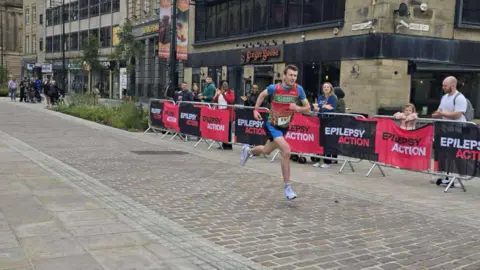 A man running in a blue and black running kit with a red vest. 