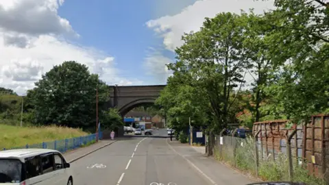 Google A long road with an arched railway bridge. There is a green space on the left and metal containers behind a metal fence on the right, as well as green trees and bushes