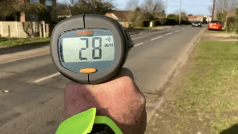 A hand holds a speed tracking device in front of a road with a car in the distance. The device has a reading of 28mph.