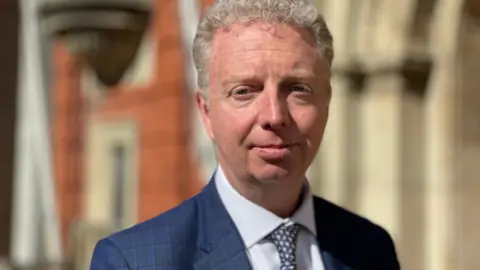 Alex Allinson is wearing a navy suit, light blue shirt and spotted tie, he has short wavy fair hair. He's standing in front of a brick building on a sunny day.