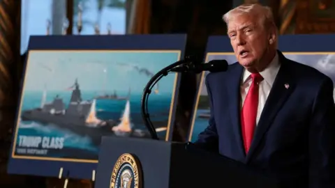 Reuters US President Donald Trump stood speaking at a lectern in a navy suit with red tie next to an image of a tanker