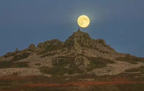 Andrew Fusek Peters A rock formation on top of a hill with a pale yellow supermoon hovering over it.