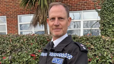 Hampshire and Isle of Wight Constabulary Chief Superintendent Simon Dodds is stood in police uniform looking at the camera. He has a light beard and is stood in front of a bush. 