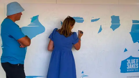 A woman uses a pen to draw on a mural, which is mostly sketched out in black and white, but some of the background has been painted blue. A man in a blue t-shirt stands next to her.