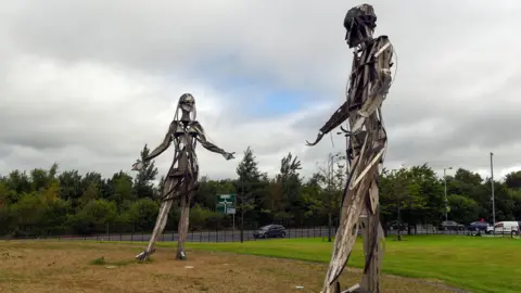 Getty Images Two semi-abstract steel and bronze statues face eachother at about 18 foot tall, with arms outstretched on a green space. Behind them is a road of traffic, backed by large trees. It is cloudy, interrupted only by a brief glimpse of blue sky in the middle of them.