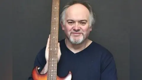 Nick Smith is looking at the camera and smiling. He has dark hair which is grey at the sides and he has a grey beard. He is holding an electric guitar and is wearing a blue pullover