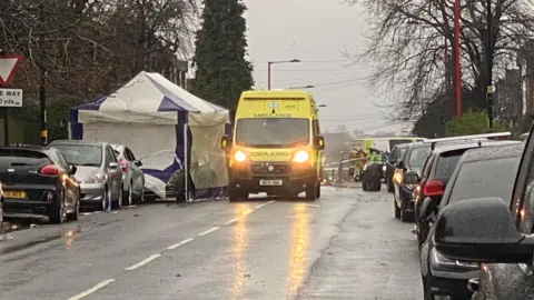 A rain-soaked road, determination   are parkland  cars either broadside  and a yellowish  ambulance with its lights on. It is adjacent  to a bluish  and achromatic  tent.