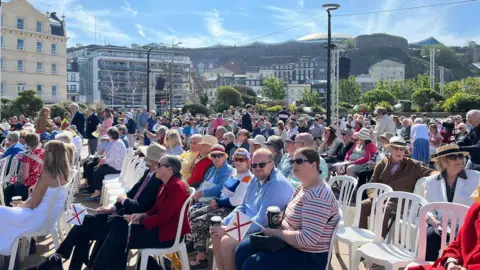 A picture of people sat down on chairs. There is a number of people in hats and holding flags
