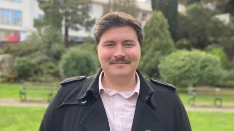 Deputy Tom Rylatt - A man with a brown quiff and a black moustache, He's wearing a black coat and a pink buttoned up shirt.