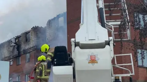 ECFRS Two firefighters operate an aerial ladder platform as they tackle the fire. The four-storey block of flats is pictured in front of them. It is an orange brick building but its top floor is totally charred and damaged by the fire.