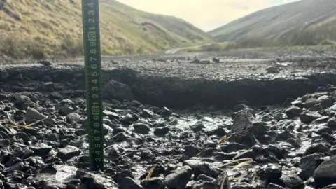 Harry Byatt A large ditch is shown with a measuring rod in it. Behind it a single track road bends around a valley.