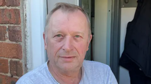 Picture of Mr Sampson stood in his doorway. He wears a grey crew neck t-shirt and a silver chain. He has blue eyes and short silver hair. He has a short stubbled beard. 