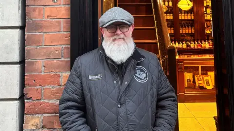 BBC A man is standing infront of a shop door. He has a black coat and grey hat on. He is wearing glasses and has a large grey beard. The building behind him is made from red bricks. Inside the shop door there is a set of steps and a shop counter. 