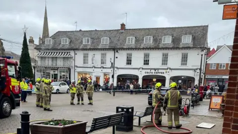 Saffron Walden Fire Station A large number of firefighters standing outside Market Place, Saffron Walden. You can see a white building, including Costa Coffee, park benches, a paved area, a Christmas tree to the left, Christmas decorations handing from buildings and a fire engine to the left. 