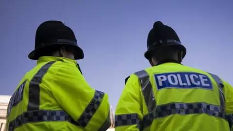 Getty Images The backs of two police officers in front of a blue sky. They are wearing neon yellow jackets that say "police" and black police hats