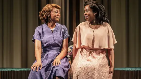 Marc Brenner Two women on stage in a theatrical production. Both are wearing hairstyles and dresses from the mid-20th Century, one dressed in purple and one salmon-coloured. They are sitting on chairs and looking at each other.