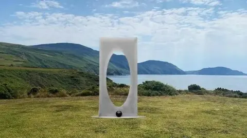 Bryan Kneale A steel modern art sculpture, which is a rectangle with an oval shaped hole, standing on some grass in front of a scenic view overlooking the sea, with coastal hills stretching into the background. 